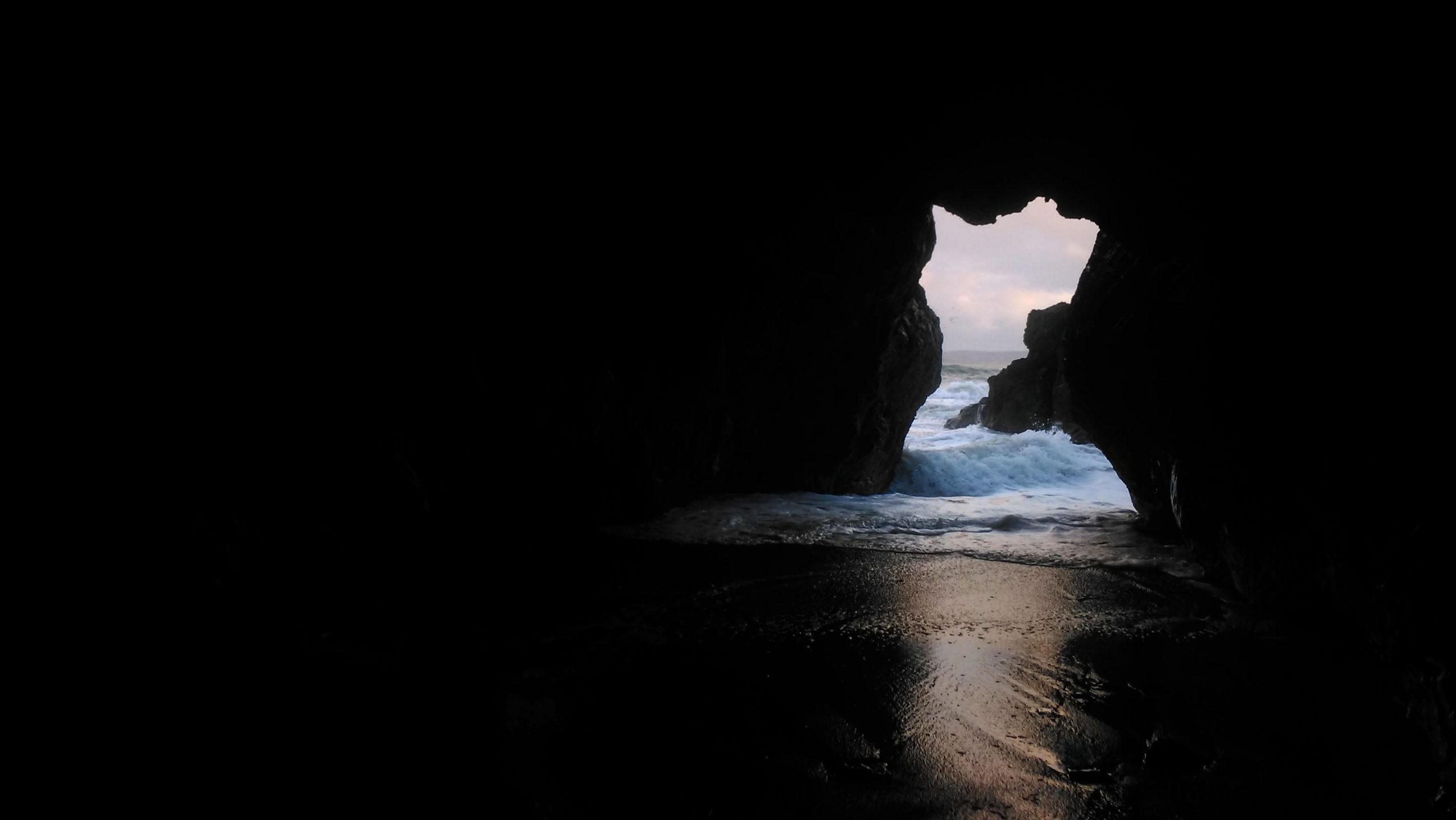 The photograph of Smugglers Cave,Portrane, Co.Dublin. The image depicts the immersiveness of being inside a cave.The opening of the cave mouth is facing outwards to the sea. The powerful tide is coming in, filling the entire entrance. The light of the sky bounces off the waves and begins to creep into the heart of the cavern.