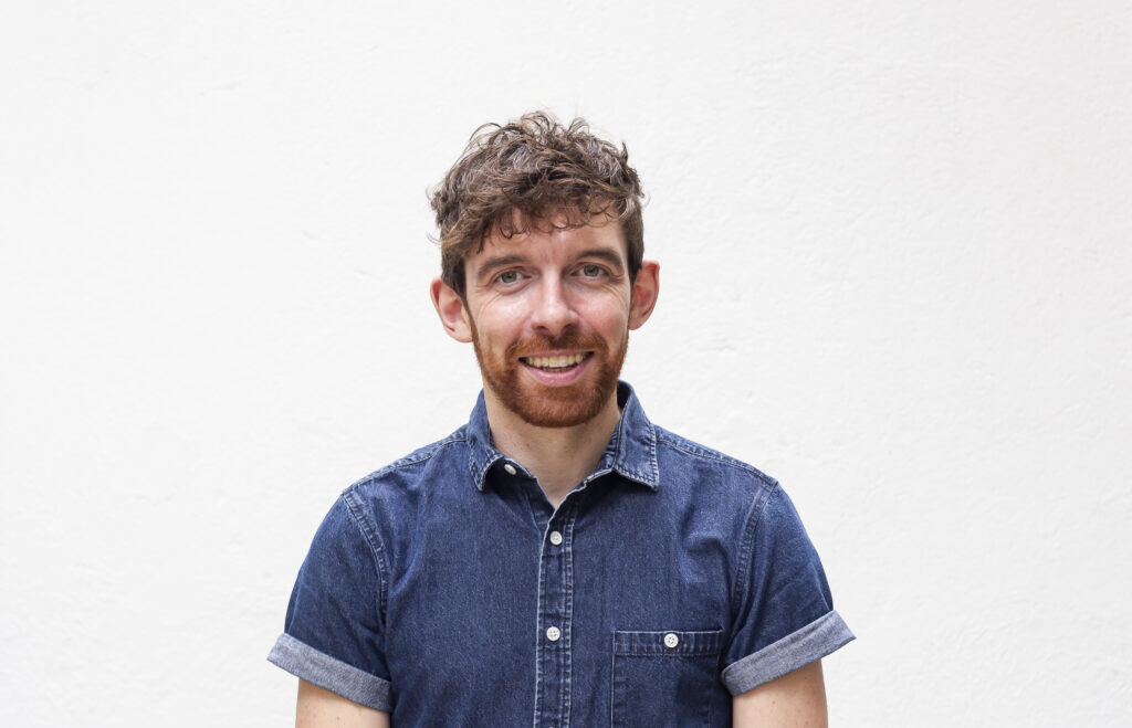 Artist AlanJames Burns, an individual with masculine features wears a deep blue denim shirt and is smiling directly towards the viewer. Their features are in a pleased and content expression, with the corners of the mouth turned up and the front teeth exposed.