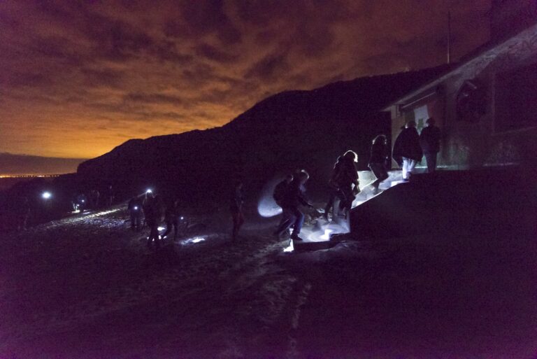A group of individuals wrapped up in winter clothing, walk in an uniformed line towards the entrance of a hut. The only light source is created by their torches held in either their hand or strapped to the head. In the background of the photograph, there is the silhouette of a mountainous landscape with the orange hint of the setting sun through the inky black clouds. A hint of the existence of a city is summoned by a faint orange glow deep in the distance.