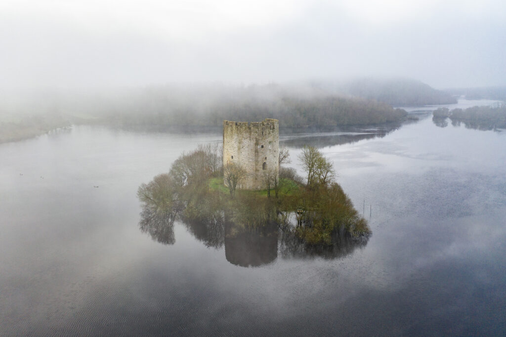 The ruin of a round stone castle town sits on a tiny island in a lake, the island is just big enough for the tower. Behind the tower the forested mainland is visible through a veil of fog. Trees surround the edge of the island and disappear into the water. The bright green grass on the island stands out in contrast to the grey of the fog and dark blue water of the lake.