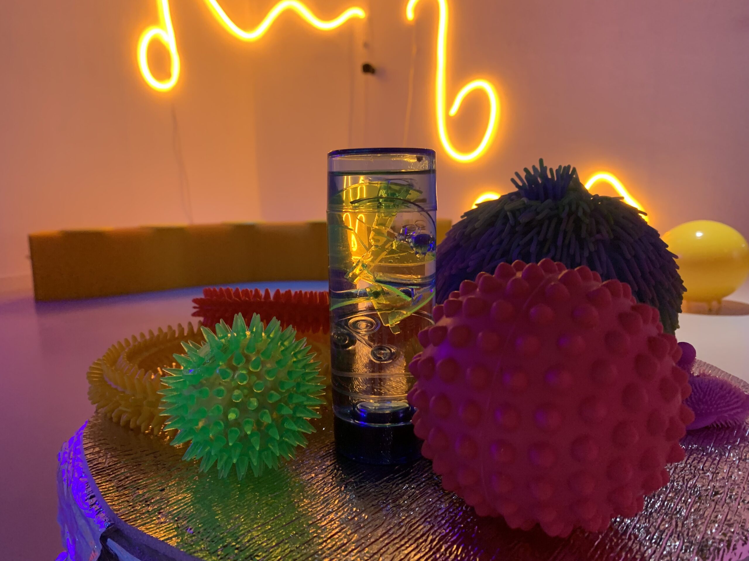 A selection of sensory objects on a table. There are balls and rings in a variety of bright colours, red, green, yellow and with a variety of textures. In the background there is a piece of looping neon art glowing orange.