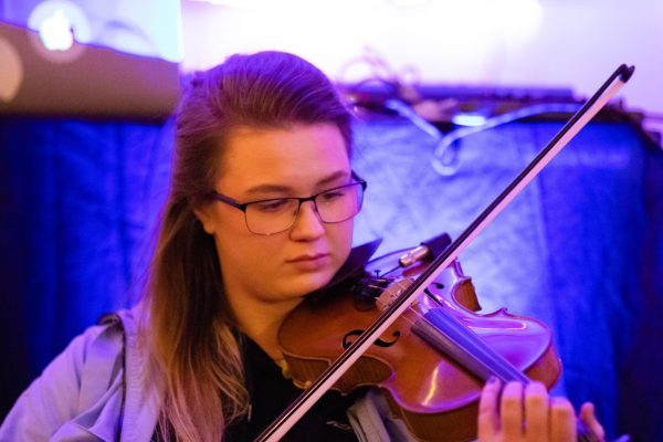 Kerry McCabe, traditional musician from Co. Fermanagh is playing the fiddle. She is wearing glasses and has long sandy coloured hair which is pulled back. There is a look of calm concentration on her face. Light hits off a blue fabric background casting an indigo light over Kerry’s face and reflecting on the surface of the fiddle.